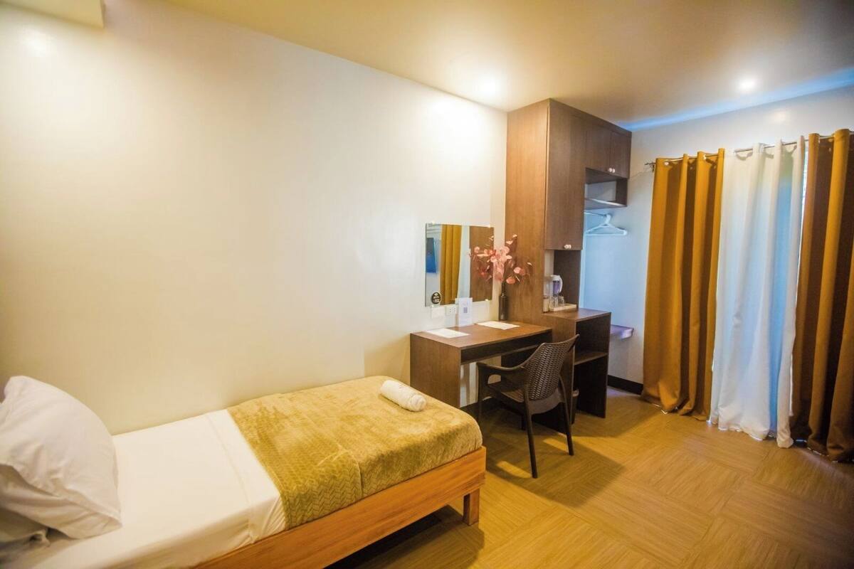 A hotel room with a single bed, a desk with a chair, a small table with a vase, and a wardrobe.