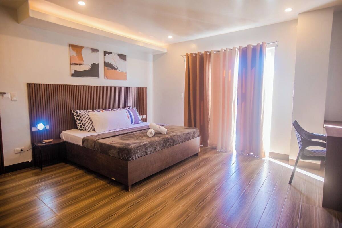 A modern bedroom with a wooden bed, a nightstand, a desk, and a large window with curtains.