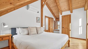 Ein Schlafzimmer mit Holzboden, einem Bett mit weißer Bettwäsche, einem Nachttisch mit Lampe und einer Tür zu einem anderen Raum.