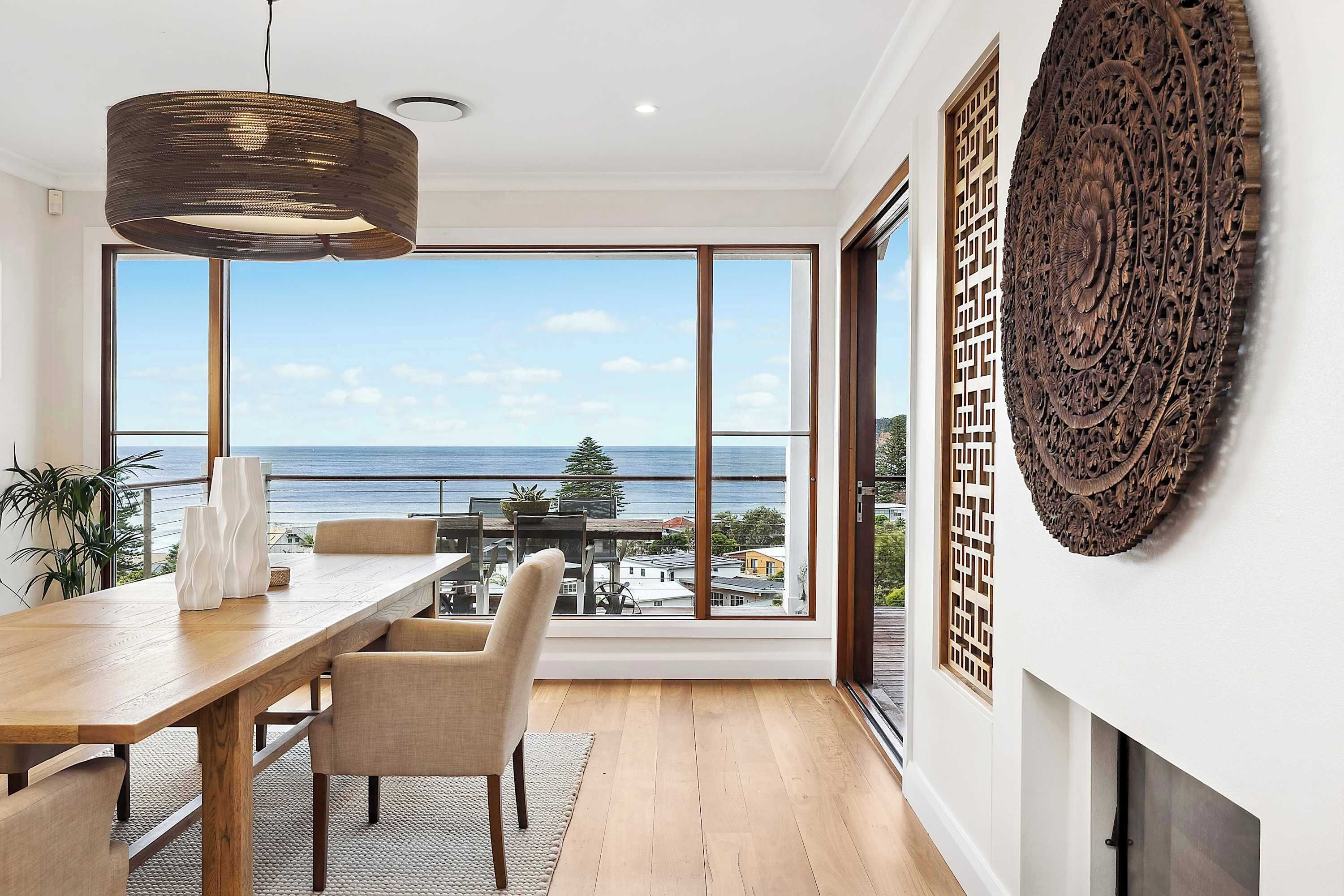 Un comedor moderno con una mesa y sillas de madera, ventanas amplias con vistas al mar y un elemento decorativo en la pared.