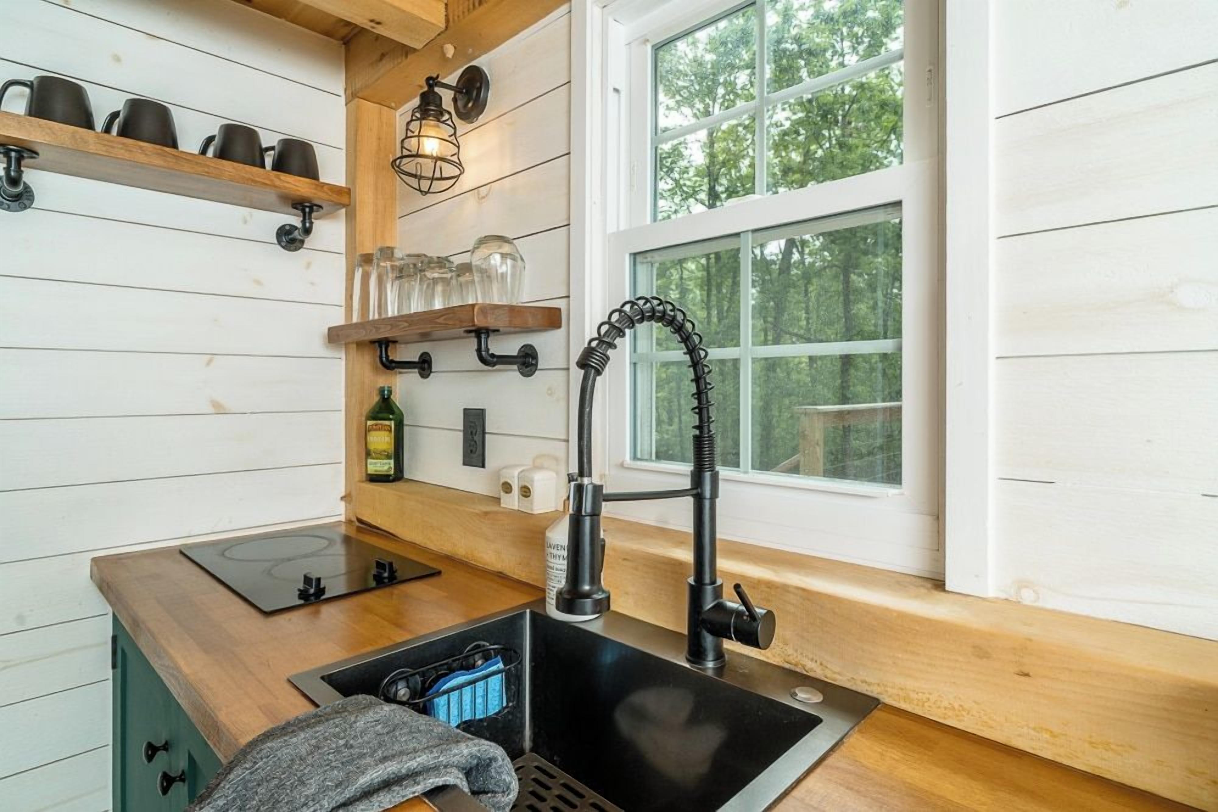 A kitchen with a black sink, wooden shelves, and a window with a view of trees.