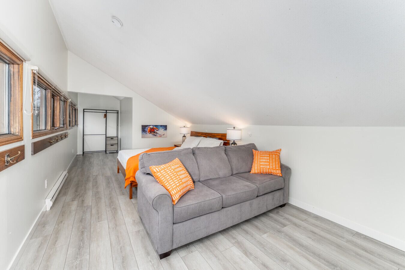 A modern, compact living space with a grey sofa, orange patterned pillows, a bed with white linens, and a wooden floor.