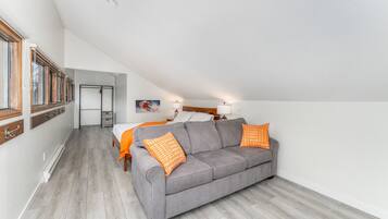 A modern, compact living space with a grey sofa, orange patterned pillows, a bed with white linens, and a wooden floor.
