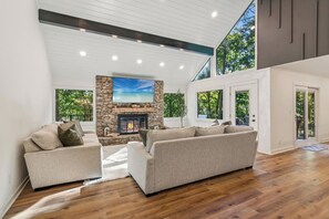 A modern living room with a stone fireplace, wooden flooring, and large windows offering a view of trees. - Modern Haven - Tree Views & Fireside Sips + Access to Pools, Trails & Golf (Jasper)