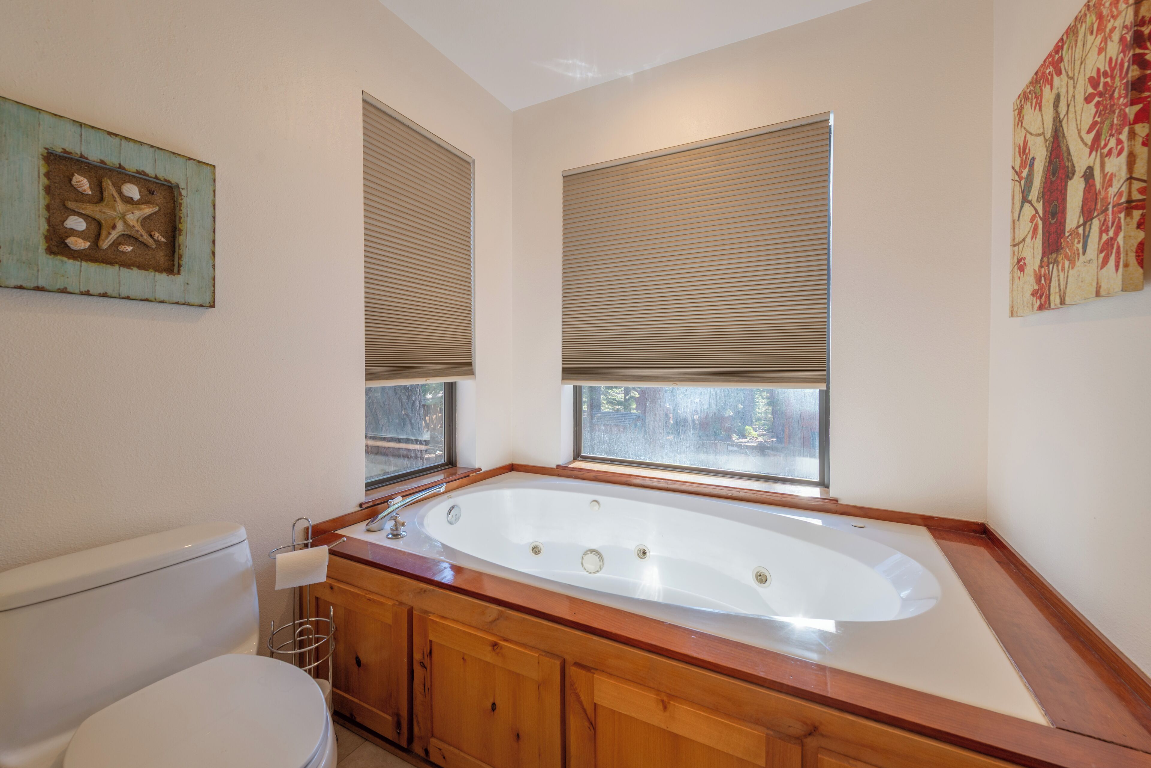 Un baño con inodoro blanco, armarios de madera, una bañera grande y dos ventanas con persianas.