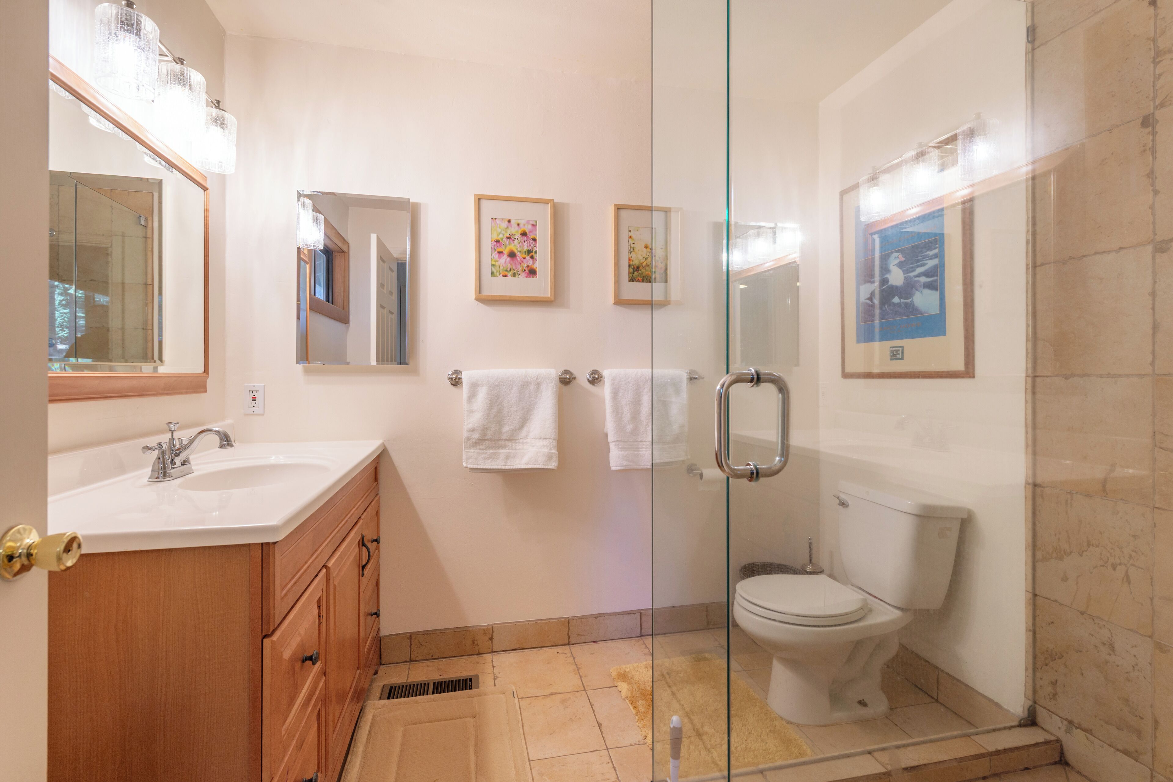 Un baño moderno con cabina de ducha de vidrio, lavamanos blanco, tocador de madera y dos cuadros en la pared.