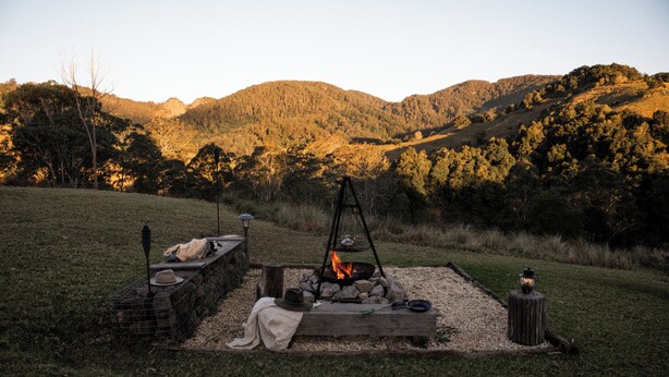 A campfire setup with a fire pit, logs, and a lantern in a grassy field with mountains in the background. - Nature Domes  (Toms Creek)