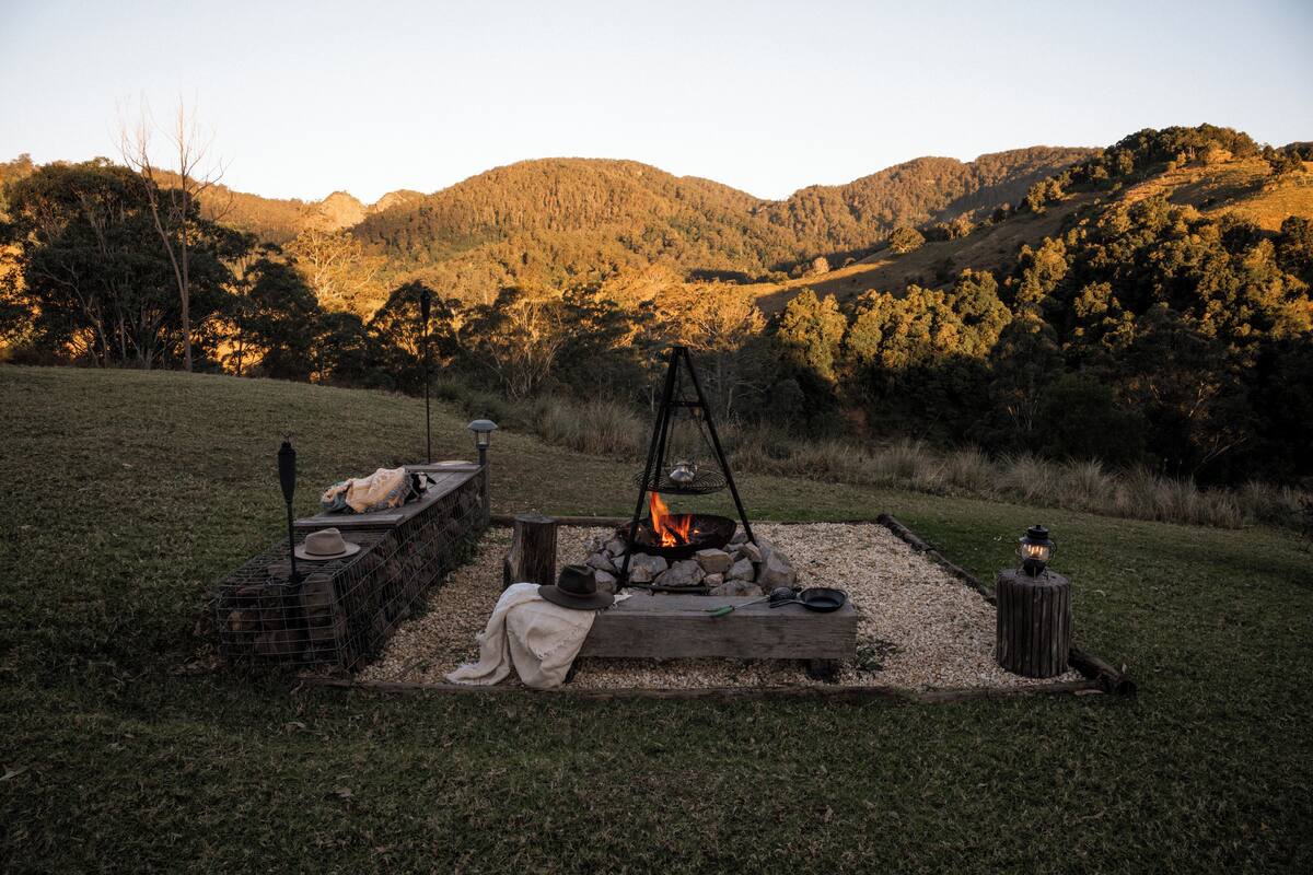 A campfire setup with a fire pit, logs, and a lantern in a grassy field with mountains in the background.