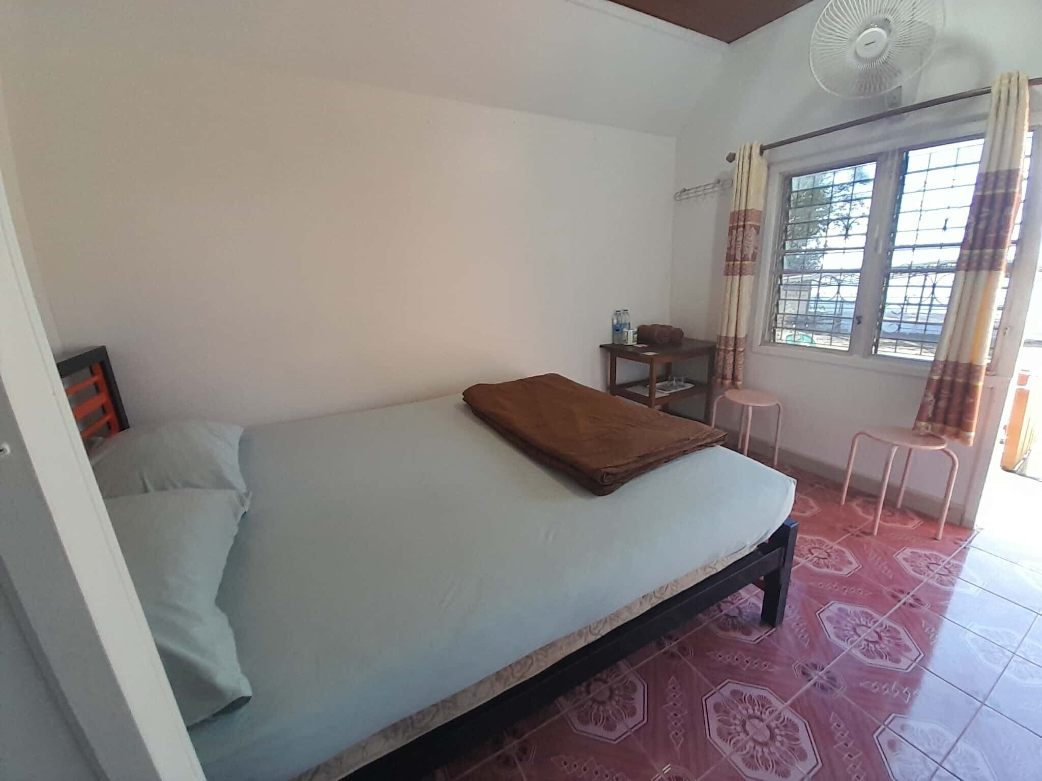 Una habitación pequeña y sencilla con cama, mesa, silla y ventana con cortinas.