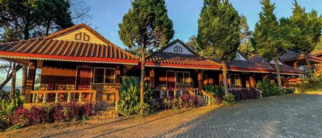 A row of traditional wooden cabins with a tiled roof, surrounded by lush greenery and trees.