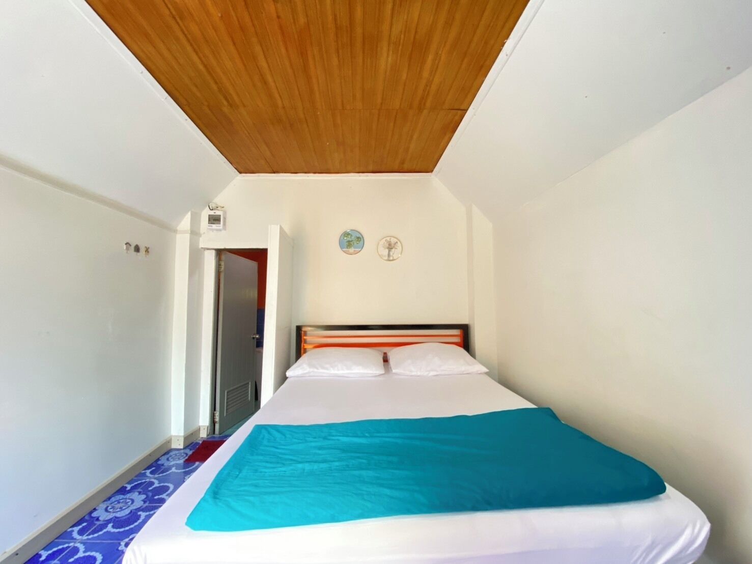 A bedroom with a wooden ceiling, a bed with white bedding and a teal blanket, a small wall clock, and a door leading to another room.
