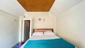Un dormitorio con techo de madera, una cama con ropa de cama blanca y una manta verde azulada, un pequeño reloj de pared y una puerta que da a otra habitación.