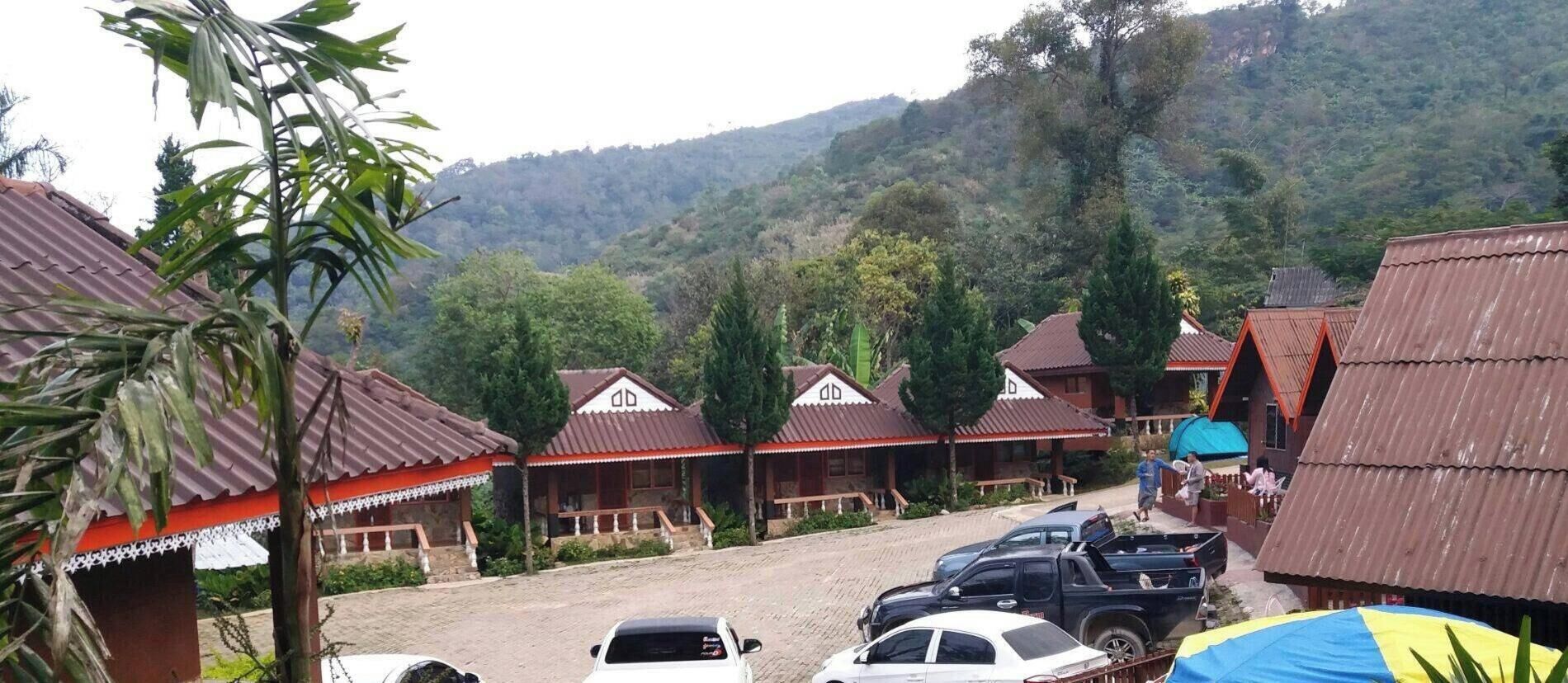 A row of traditional-style buildings with red roofs and white trim, set against a backdrop of lush greenery and hills.