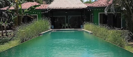 A swimming pool with a tiled edge, surrounded by greenery and a building with a thatched roof.