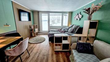 Ein modernes, gut beleuchtetes Zimmer mit Holzboden, einem Bett mit dunklem Kopfteil, einer Couch, einem kleinen Tisch und einem an der Wand montierten Fernseher.