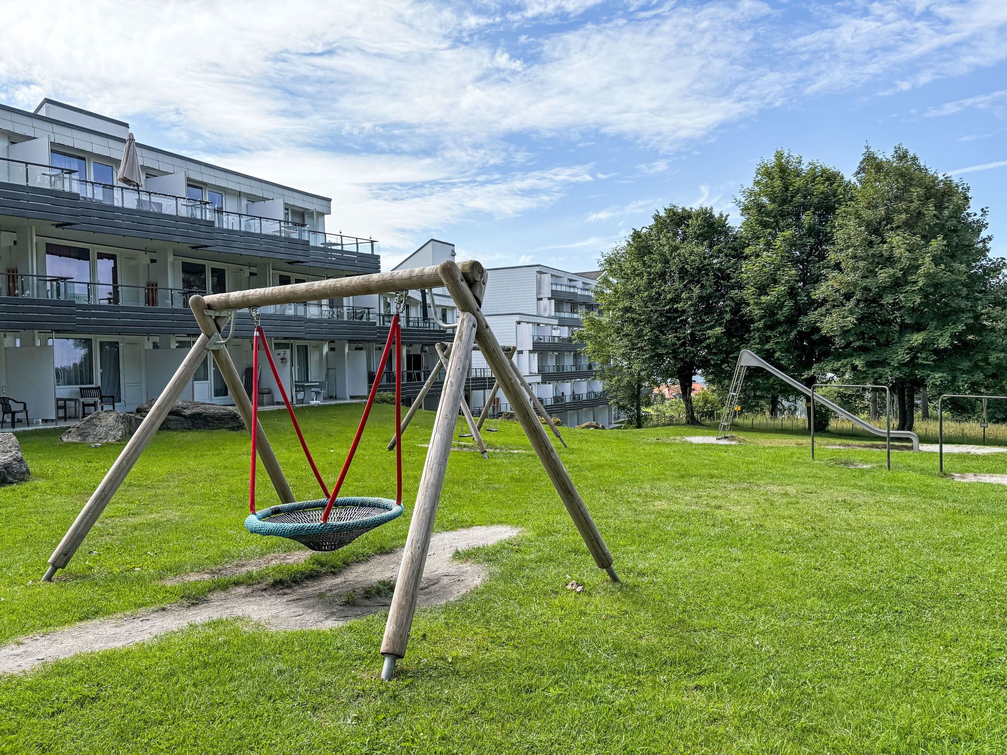 A playground with swings and a slide, surrounded by grass and trees, with buildings in the background.