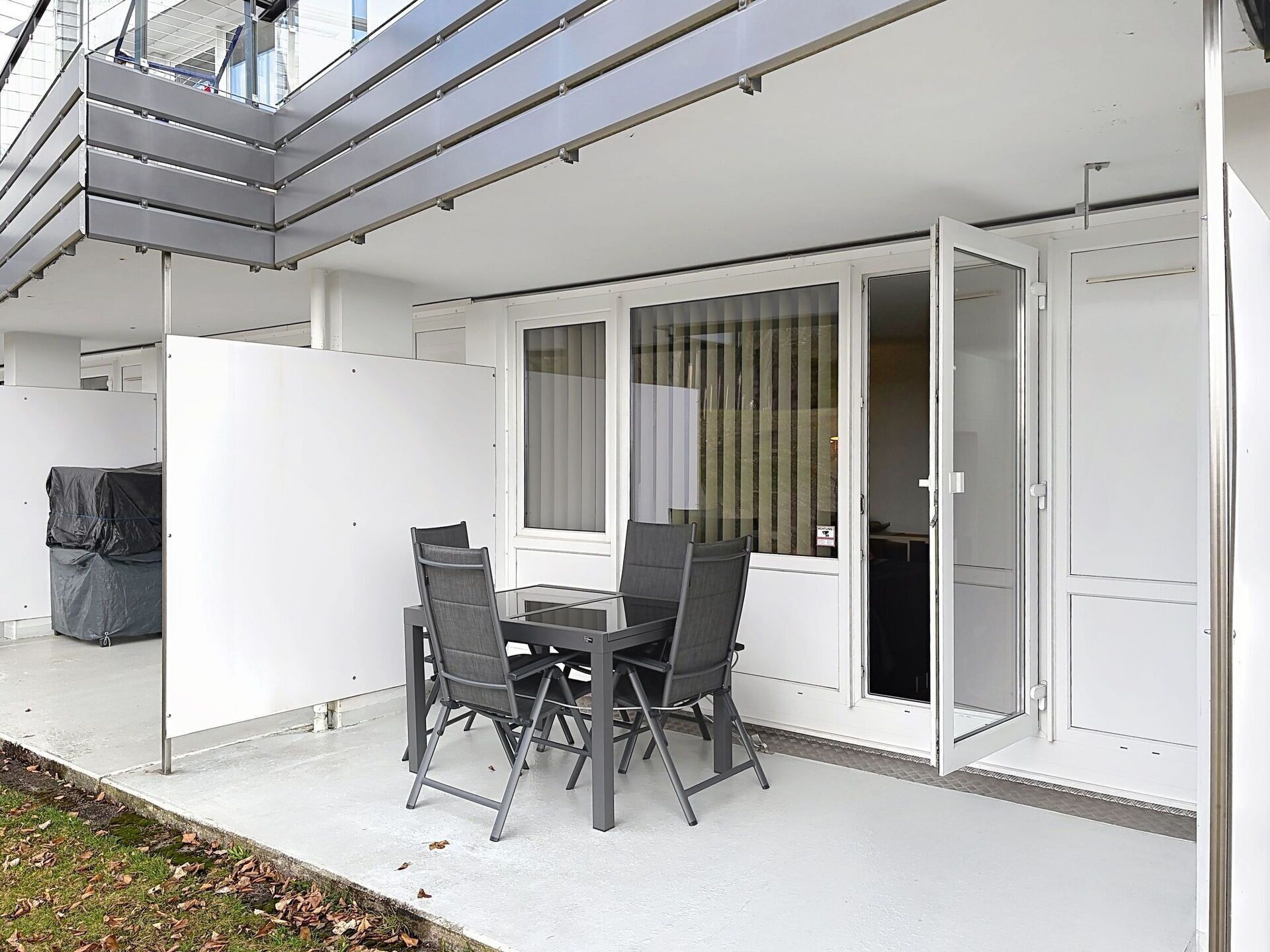 An outdoor patio with a glass table and chairs, a sliding glass door, and a covered grill area.