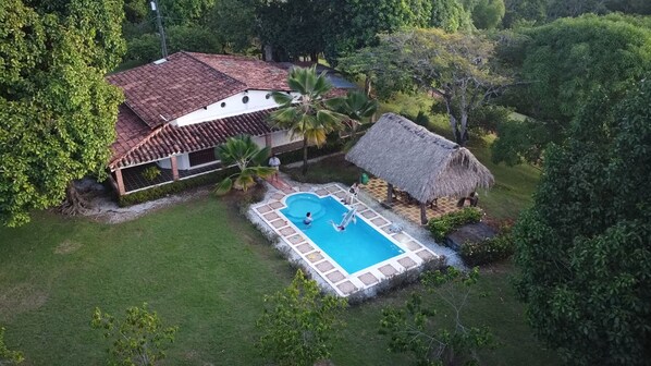 Vista aérea de una casa con piscina, rodeada de exuberante vegetación y una construcción con techo de paja.