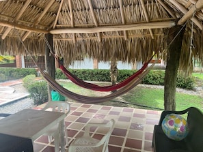 Una hamaca bajo un techo de paja, una mesa, sillas y vistas a un jardín ajardinado.