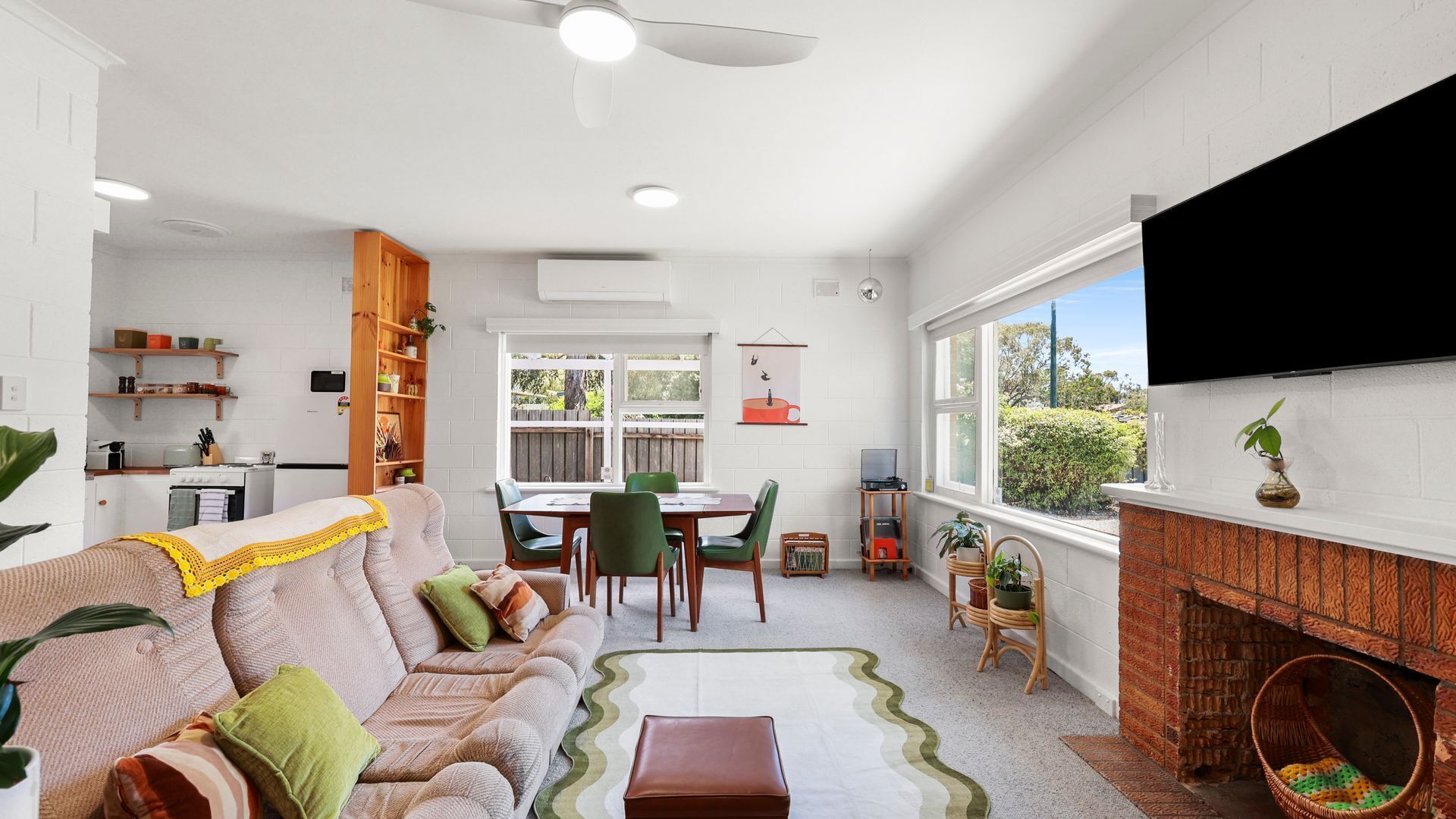 A modern living room with a sofa, coffee table, and dining area. There is a kitchen with a stove and shelves. A TV is mounted on the wall, and there are plants and a fireplace.