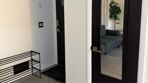 A modern hallway with a black door, a welcome sign, a black metal rack, and a glimpse of a living room with a sofa and a plant. - Modern Fully Renovated 2BR Retreat • Minutes from RIT • Safe & Spacious (Rochester)