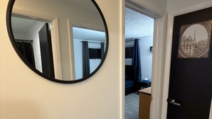 A hallway with a round mirror on the left, a door with a picture frame on the right, and a glimpse of a bedroom with a bed and curtains. - Modern Fully Renovated 2BR Retreat • Minutes from RIT • Safe & Spacious (Rochester)