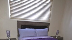 Een slaapkamer met een bed, twee nachtkastjes, een raam met jaloezieën en een nachtlampje op elk nachtkastje.
