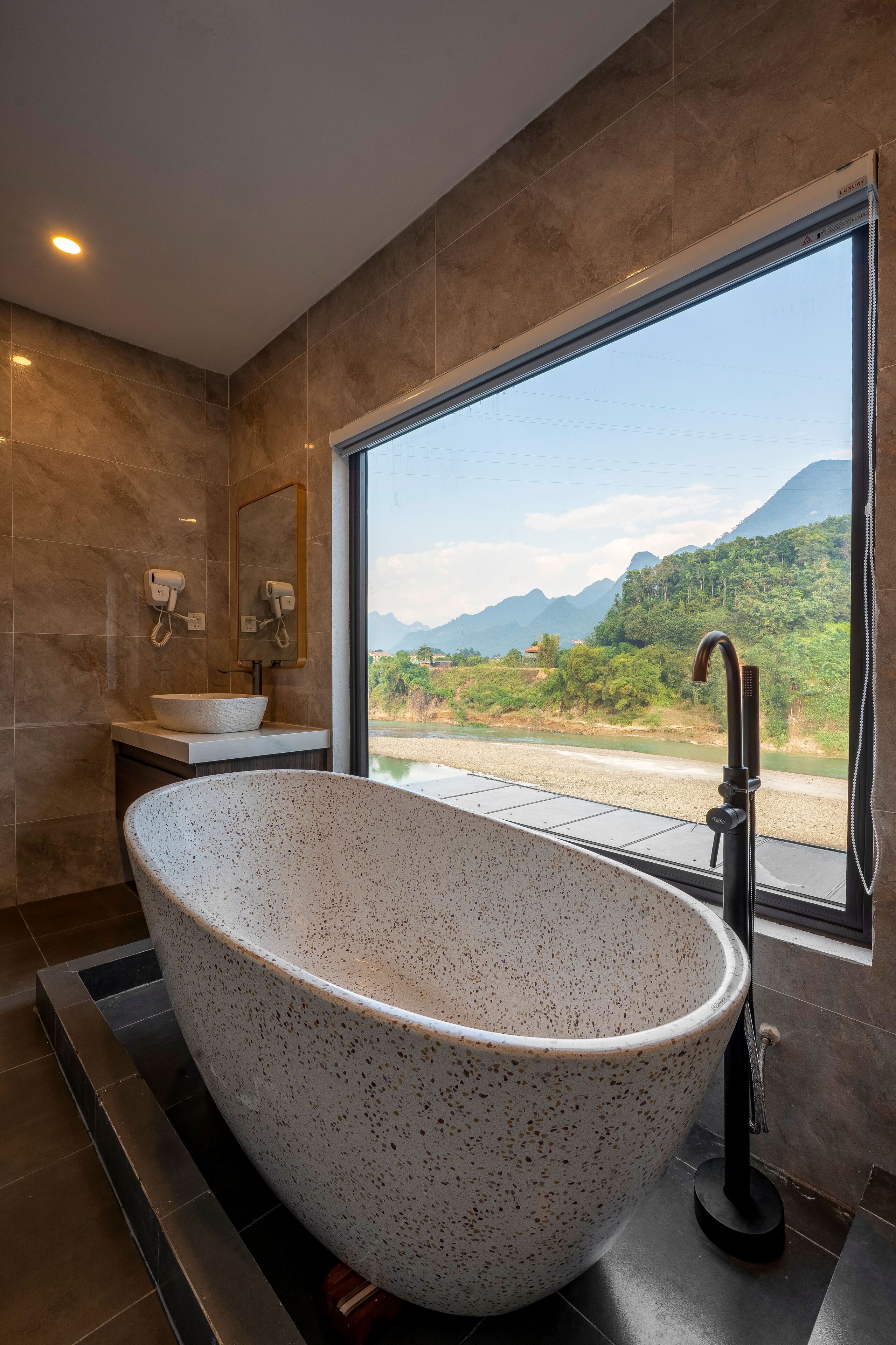A modern bathroom with a freestanding bathtub, a sink, and a large window offering a view of a river and mountains.