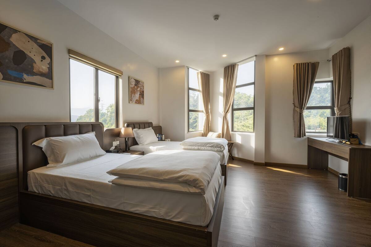 A hotel room with two beds, a desk, and large windows.