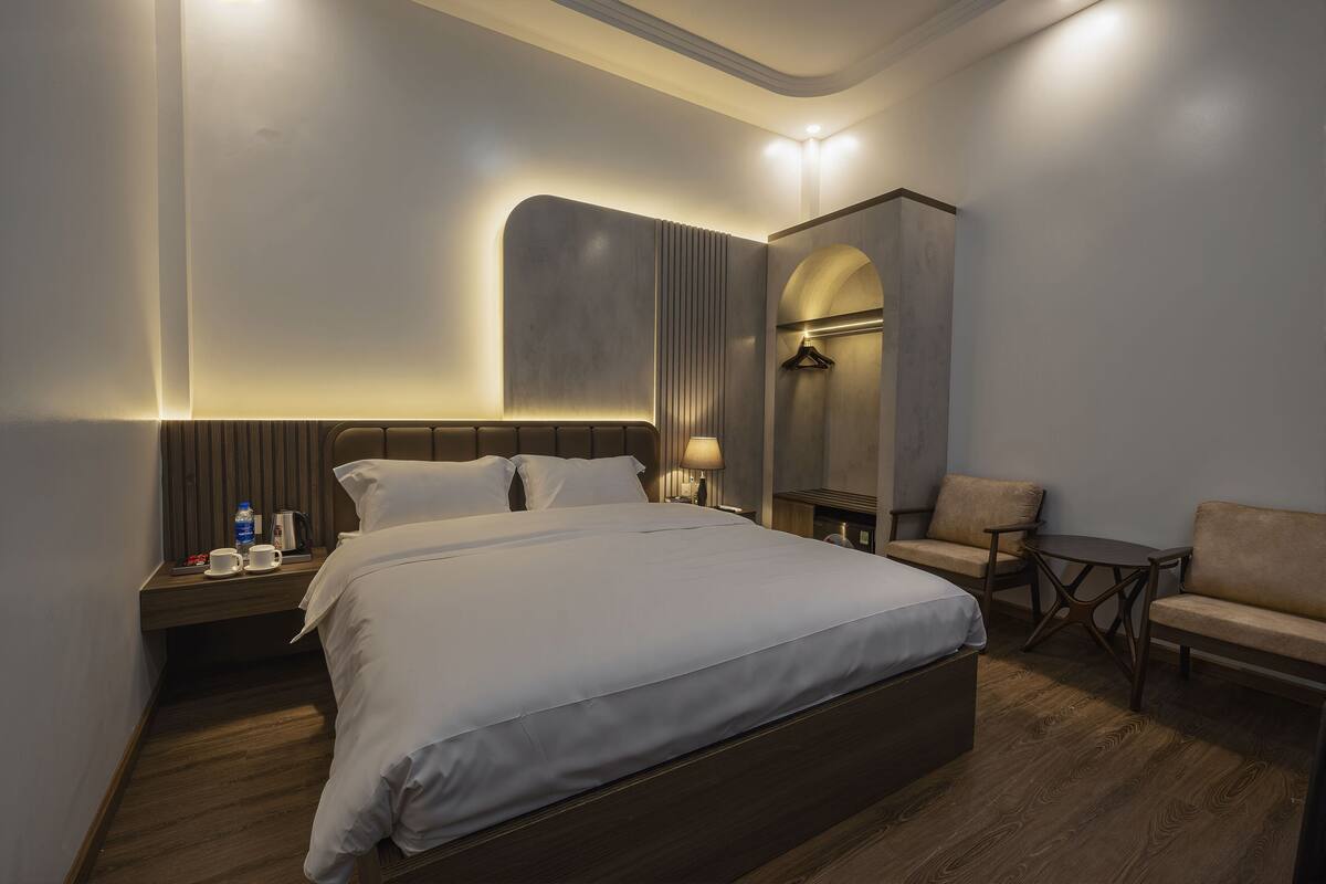 A modern hotel room with a large bed, a small table, a chair, and a wardrobe.