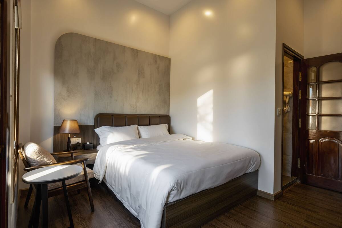 A bedroom with a large bed, a bedside table with a lamp, a chair, and a wooden door.