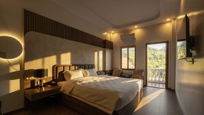 A modern hotel room with a large bed, bedside lamps, a nightstand, a chair, and a balcony with a view of greenery. - Odyssey Hotel 2 (Ha Giang 1)