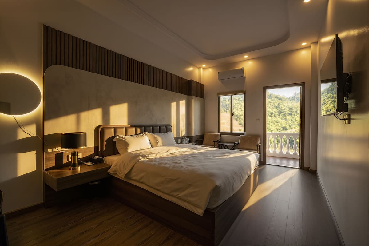 A modern hotel room with a large bed, bedside lamps, a nightstand, a chair, and a balcony with a view of greenery.