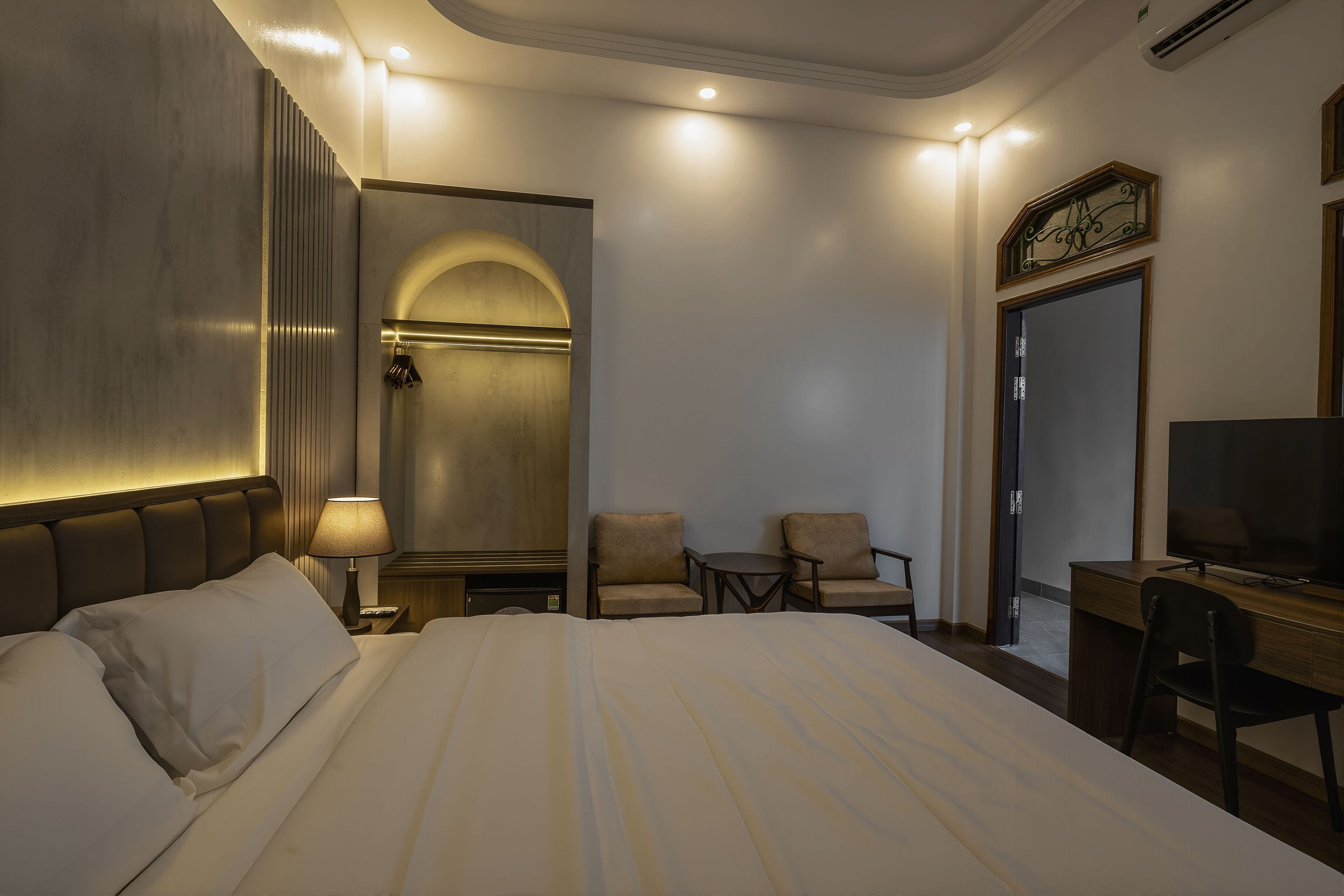 A hotel room with a large bed, two armchairs, a desk with a TV, and a wardrobe.