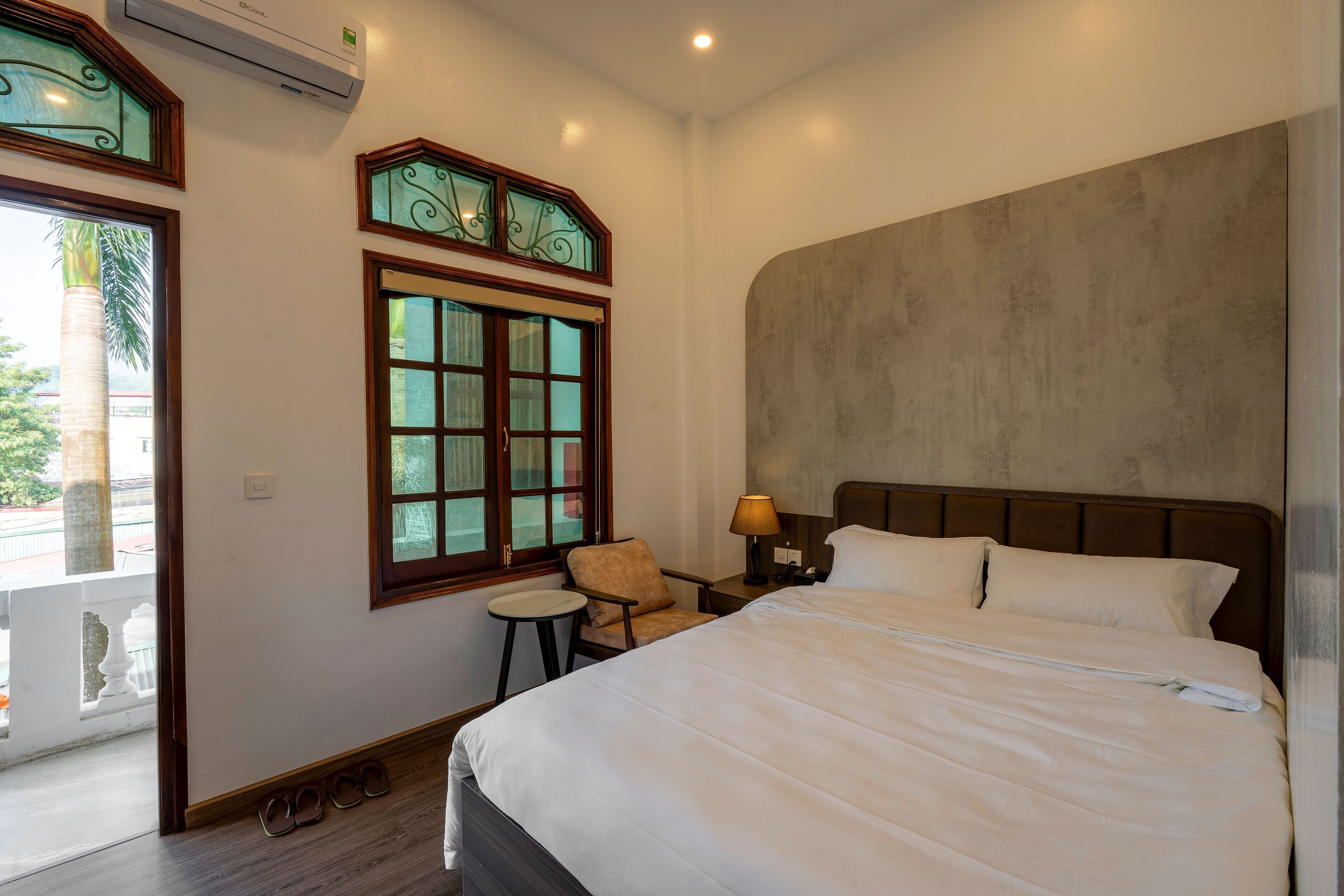 Quarto de hotel com uma cama grande, uma mesa pequena, uma cadeira, uma mesa de cabeceira com abajur e uma janela com vitrais decorativos.