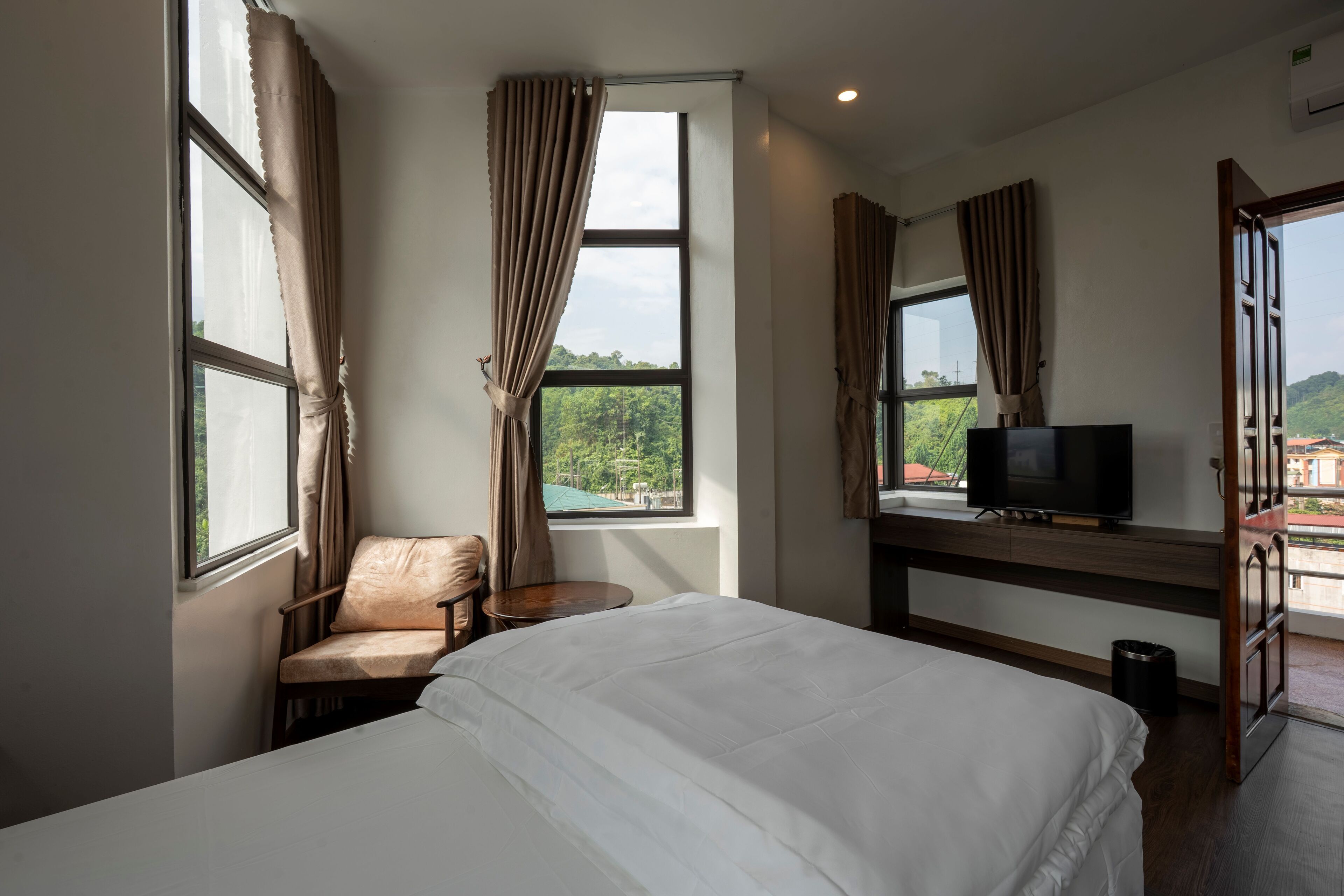 A hotel room with a bed, a chair, a small table, a TV, and a window with curtains.