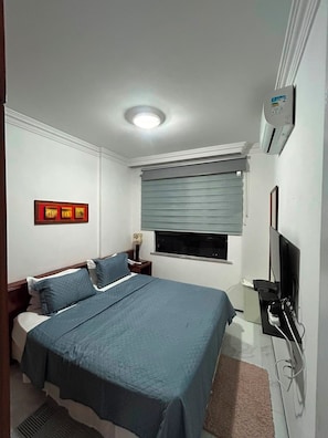 Een slaapkamer met een bed, nachtkastje, lamp, televisie en een raam met zonwering.