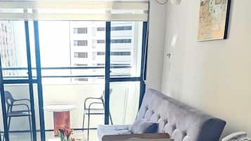 Uma sala de estar com um sofá cinza, uma mesinha redonda com um vaso de flores, uma mesa de centro de vidro e uma mesa auxiliar de vime com um telefone.