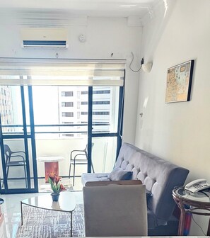 Uma sala de estar com um sofá cinza, uma mesinha redonda com um vaso de flores, uma mesa de centro de vidro e uma mesa auxiliar de vime com um telefone.