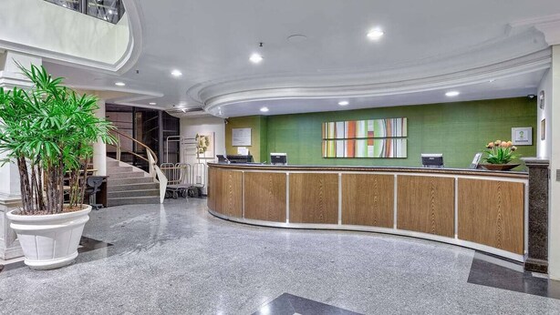A reception area with a curved wooden counter, a green accent wall, and a staircase in the background. - Beira Mar Apartment - Fortaleza (Fortaleza)