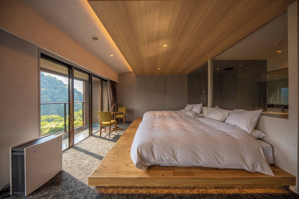 Une chambre moderne avec un grand lit, une plateforme en bois, une vue sur la verdure et une porte-fenêtre coulissante.