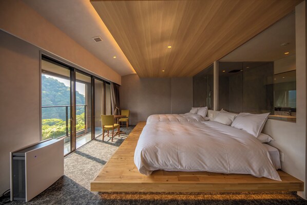 A modern bedroom with a large bed, a wooden platform, a view of greenery, and a sliding glass door. - THE ACONCAGUA RESORTS (Minamiaso)