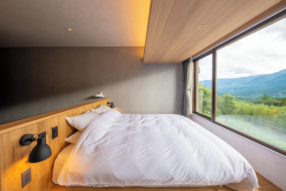 Une chambre moderne avec une grande fenêtre, une tête de lit en bois et un lit recouvert de linge de lit blanc.