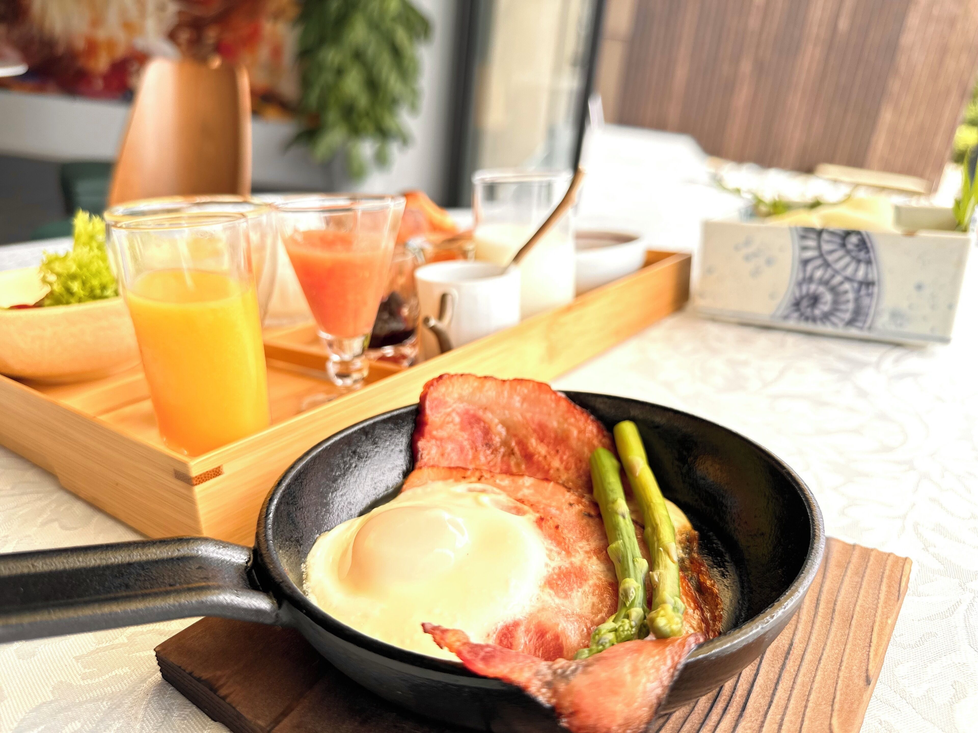 A breakfast tray with a skillet containing bacon, an egg, and asparagus, accompanied by glasses of orange juice and a smoothie.