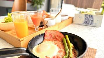 Bandeja de desayuno con una sartén que contiene tocino, un huevo y espárragos, acompañada de vasos de jugo de naranja y un batido.