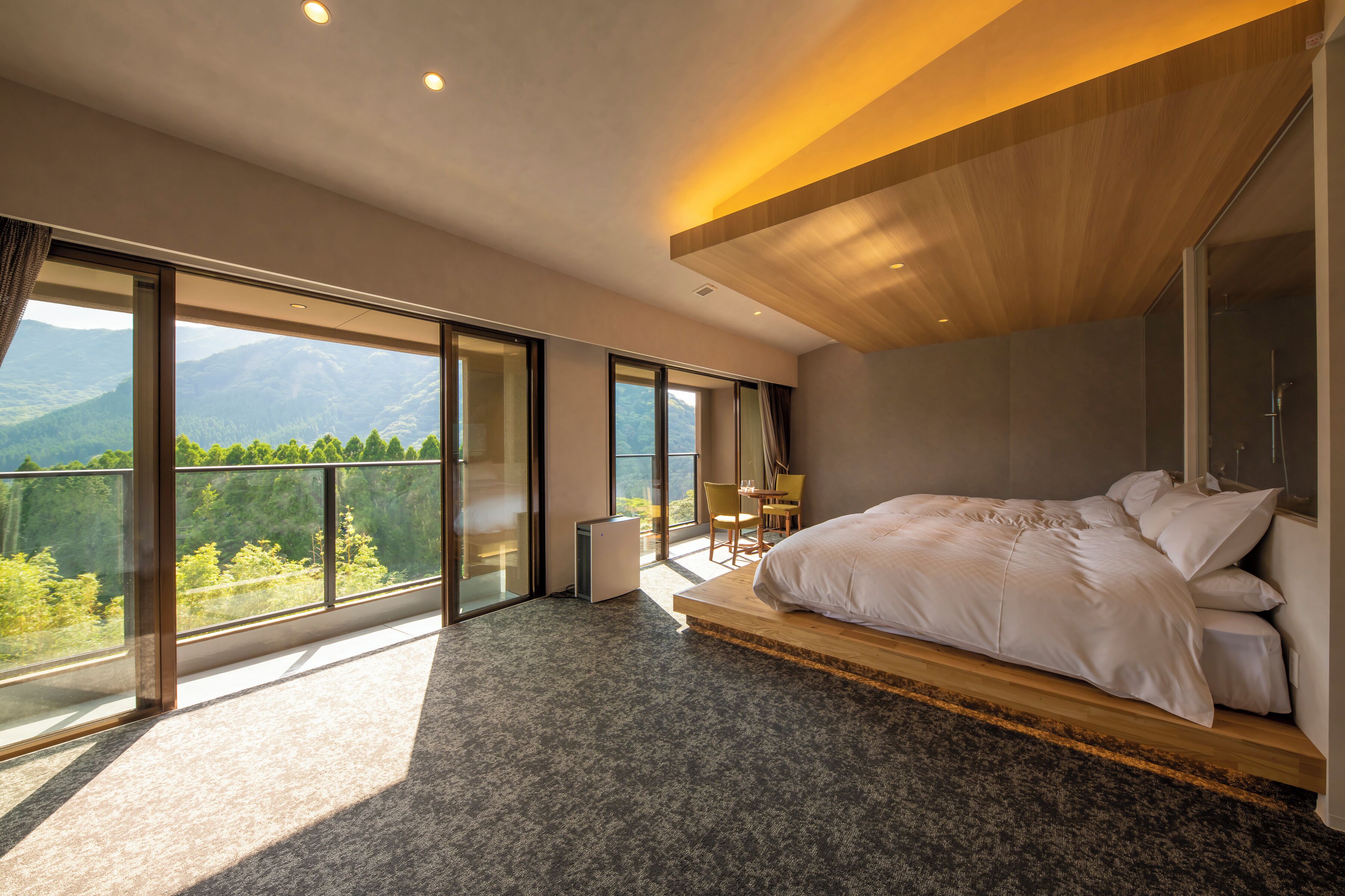 A modern bedroom with a large bed, a small table, and a chair, large windows with a mountain view, and a wooden ceiling.