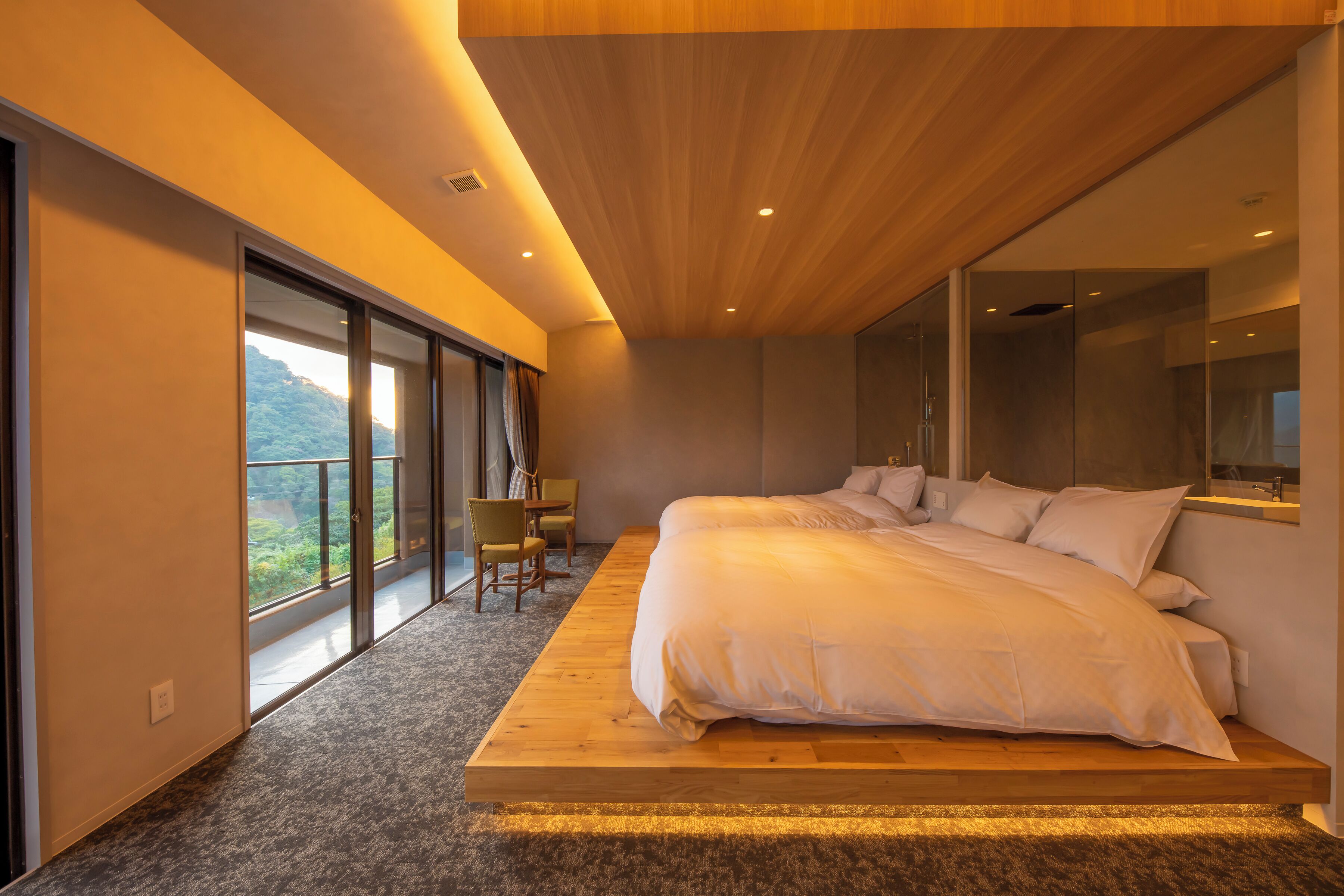 A modern bedroom with a large bed, a wooden platform, a seating area with chairs, and a view of the outdoors through large windows.