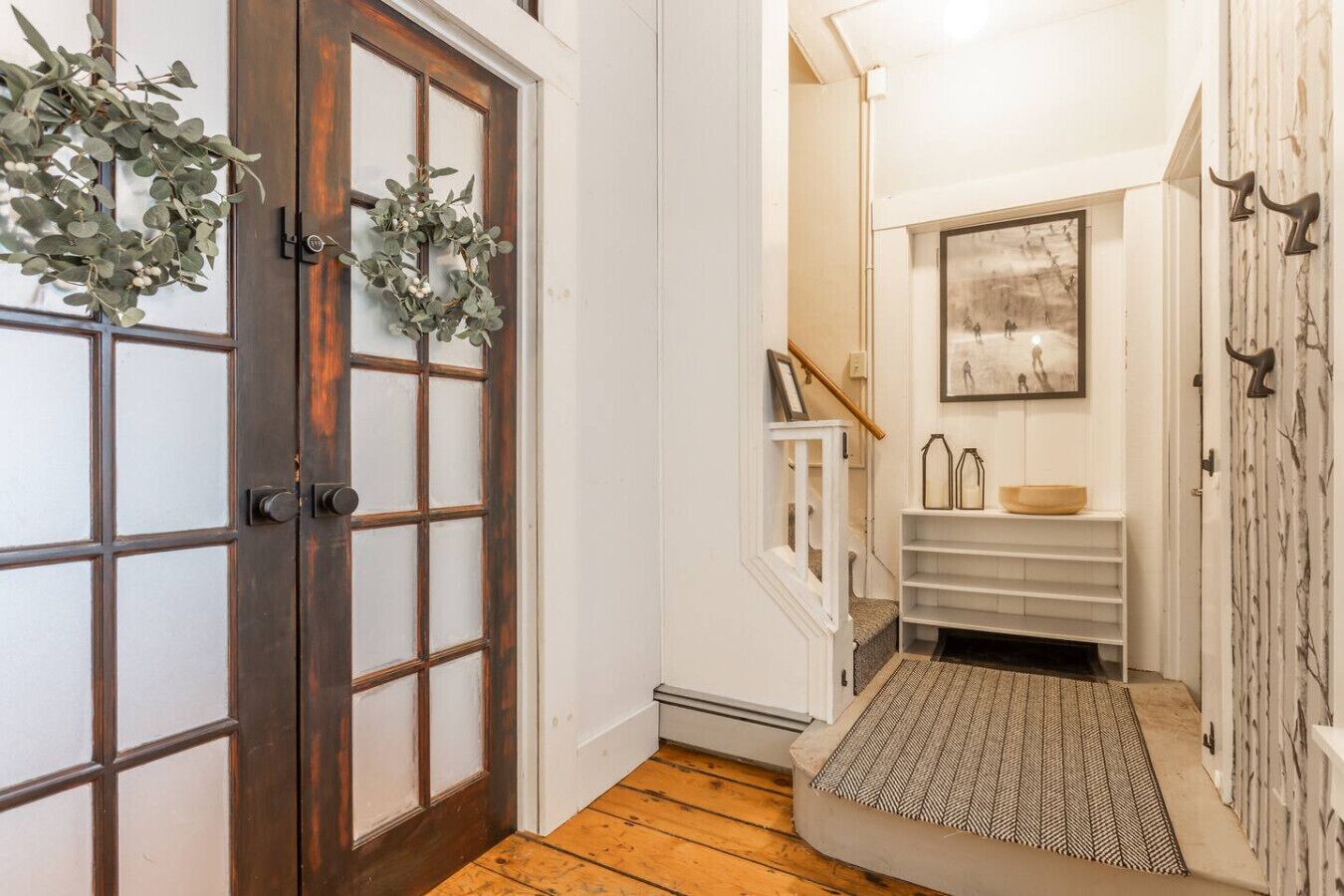 Um corredor com porta de madeira, parede branca, escada, quadro emoldurado e um pequeno prateleira com objetos decorativos.