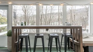 A room with a wooden table and three black stools, a window with a view of trees, and string lights. - Ski Oasis w/ Hot Tub near Killington & Woodstock (Bridgewater)