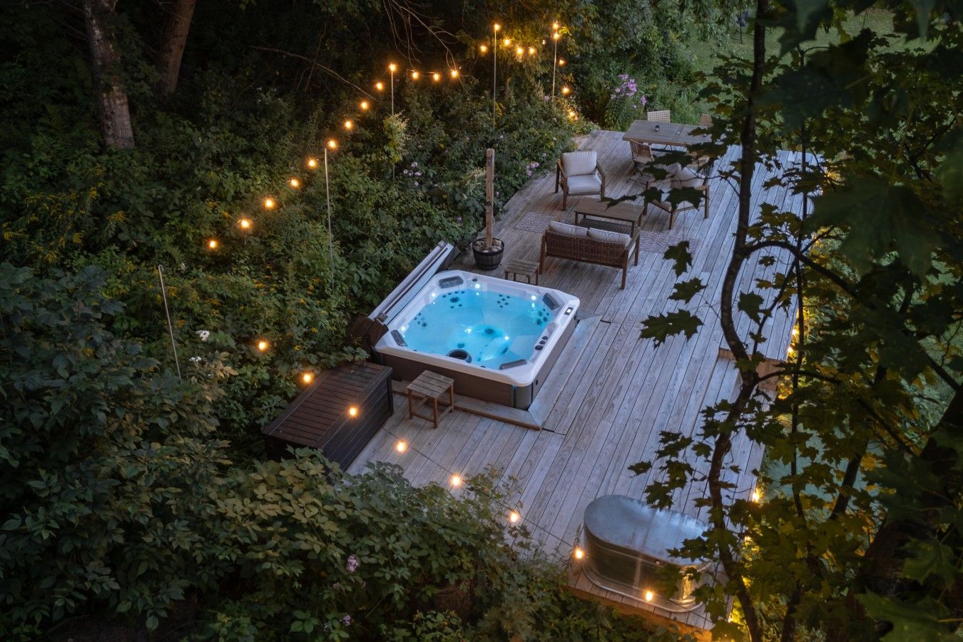 Un bain à remous entouré de meubles de jardin en bois, de guirlandes lumineuses et d’une végétation luxuriante.